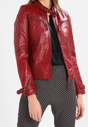 Veste en cuir rouge avec poches zippées sur la poitrine et sangles à boucles aux manches portée sur un haut noir et un pantalon à motifs.