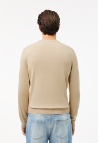 Mann mit braunen Haaren, der einen beigen Langarmpullover und hellblau gewaschene Jeans trägt, steht mit dem Rücken zu einem einfachen weißen Hintergrund.