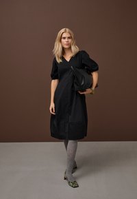 Ženska v črni midi obleki in sivih nogavicah drži veliko črno teksturirano torbico, stoji ob rjavi steni in sivi tleh.