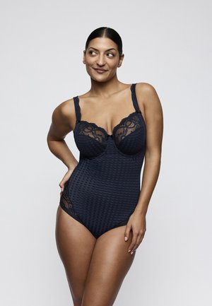 Woman with dark hair in a low bun wearing a black lace-trimmed bodysuit, standing with one hand on her hip and looking to the side.