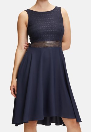 Mouwloze marineblauwe jurk met kanten gestructureerd lijfje en doorschijnend taillepaneel, uitlopende rok die boven de knieën valt, gedragen door een staande persoon.