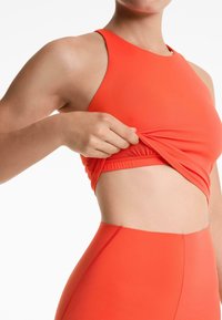 Fényes narancssárga sportos cropped top, illeszkedő fazonnal, állítható pántokkal és redőzött anyaggal, párosítva illeszkedő magas derekú leggingssel.