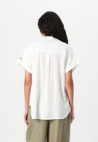 Camicia bianca a maniche corte con vestibilità rilassata, dotata di colletto, risvolti con bottoni dorati e una cucitura centrale sul retro.