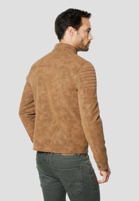Veste en daim marron à texture douce, avec des accents côtelés aux épaules et un col simple. Portée avec un jean en denim foncé.