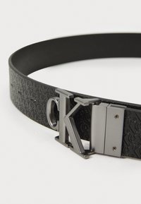 Zwarte leren riem met een reliëf logo patroon, een metalen gesp met een glanzende zilveren afwerking en een strak, modern ontwerp.