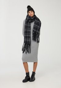 Sciarpa a quadri in bianco e grigio con bordi frangiati, indossata sopra un vestito grigio a costine. Cappellino di maglia scura con un dettaglio a pom-pom, abbinato a stivali neri.