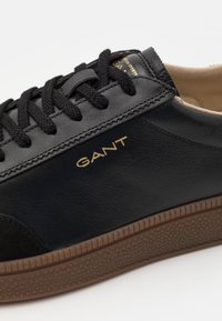 Schwarze Leder-Sneaker mit Wildlederakzenten, strukturiertem braunem Gummisohle und schwarzen Schnürsenkeln. „GANT“-Logo in Gold auf der Seite geprägt.