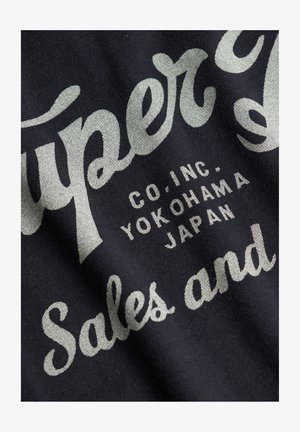 Tissu noir avec texte vintage blanc et gris comprenant "Super" et "Ventes et" ainsi que des informations supplémentaires sur l'entreprise.