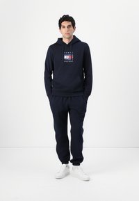 Haut à capuche bleu marine avec cordon de serrage, poche avant et logo ; associé à un jogging assorti et des baskets blanches. Tissu doux avec des poignets côtelés.