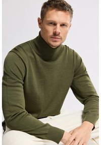 Homme avec des cheveux châtain clair courts portant un pull à col roulé vert olive et un pantalon crème assis les mains croisées.