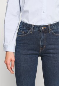 Persona que lleva unos pantalones de mezclilla azul oscuro y una camisa de botones azul clara, con la mano izquierda relajada a su lado.