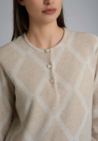 Donna che indossa un maglione beige con leggere diagonali a motivo romboidale e tre bottoni rivestiti di tessuto vicino al colletto.