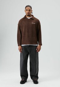 Pull-over en polaire marron avec un col, arborant un logo blanc. Associé à un jean ample foncé et des chaussures noires à lacets.