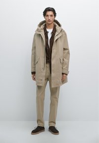 Beige parkas med huva, med dragkedjestängning och framfickor, lager på en brun skjorta och krämfärgade byxor, kompletterat med mörka skor.