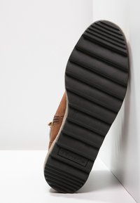 Brązowy zamszowy but z czarną gumową podeszwą, z rowkami zapewniającymi przyczepność, bocznym zamkiem błyskawicznym oraz subtelnym logo na podeszwie.