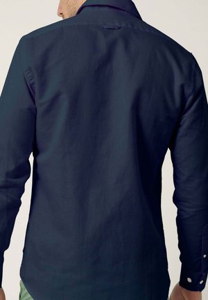 Hombre con una camisa de manga larga de color azul marino oscuro, mostrado de espaldas, de pie contra un fondo claro y liso.