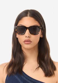 Ochelari de soare cu ramă pătrată în stil tartă, lentile închise la culoare și brațe proeminente negre. Designul include accente subtile și un finisaj lucios.