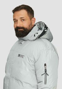 Beverde man in een lichtgrijze gewatteerde jacks met capuchon, zwarte ritszak op de mouw en het logo "Elevated Casual Est. 1969" op de borst.