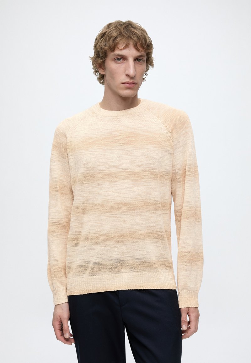 Jeune homme aux cheveux blonds bouclés portant un pull rayé beige et marron clair ainsi qu'un pantalon sombre, debout devant un fond uni.