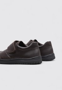 Zapatos de cuero marrón con punta redondeada, correa de velcro y suela de goma texturizada. Diseño simple con costuras y acentos mínimos.