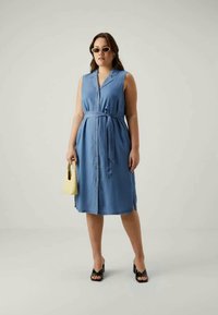 Lyseblå, ermeløs denimkjole med knappfront og knyttet midje. Strukturert stoff, knelengde med sideslits. Båret med svarte hæler og en gul veske.