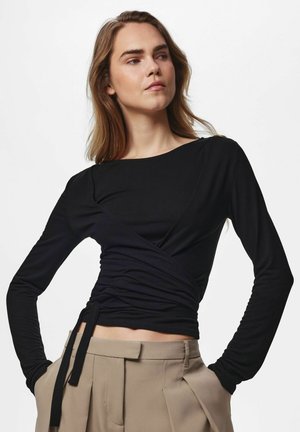Femme aux longs cheveux châtain clair portant un haut noir froncé à manches longues et un pantalon beige taille haute les mains dans les poches.