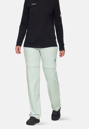 Pantalons zippés vert clair en tissu léger, arborant un logo sur la jambe gauche, associés à un haut noir à manches longues.