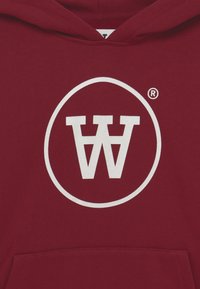 Bordeaux hoodie met een wit rond logo in het midden, met elkaar overlappende letters "W" en "A". Materiaal is zacht en getextureerd.