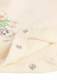 Teren de bumbac crem cu un guler texturat și ribat. Prezentă butoane metalice argintii și un design imprimat cu flori și un personaj de desene animate.
