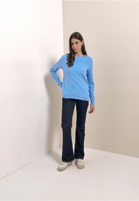 Pull bleu avec une finition texturée, col rond et poignets côtelés, assorti à un jean flare foncé et des baskets blanches. Fond uni.
