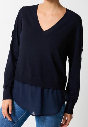 Personne portant un pull bleu marine à col en V superposé à un chemisier bleu marine transparent, associé à un jean bleu, montré de la poitrine aux hanches.
