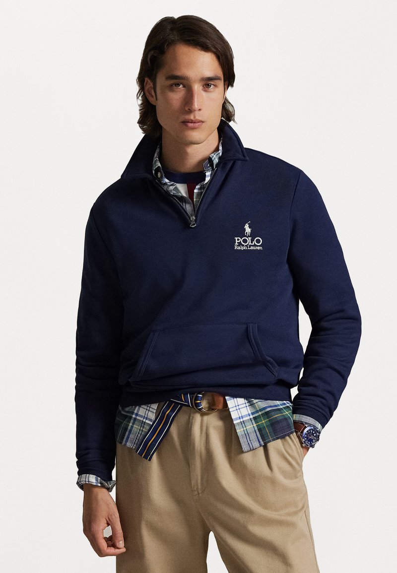 Sweatshirt azul marinho com gola de meia-zip, apresentando um bolso frontal e logótipo bordado. Usado sobre uma camisa às linhas e calças bege.
