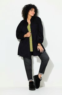 Camicia oversize in corduroy nero, top a fantasia verde, jeans scuri e stivaletti neri con superficie testurizzata. I capelli ricci aggiungono volume.