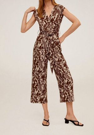 Kvinde iført beige og brun blomsterprintet kortærmet jumpsuit med vide ben, bindebælte og sorte hælsandaler.
