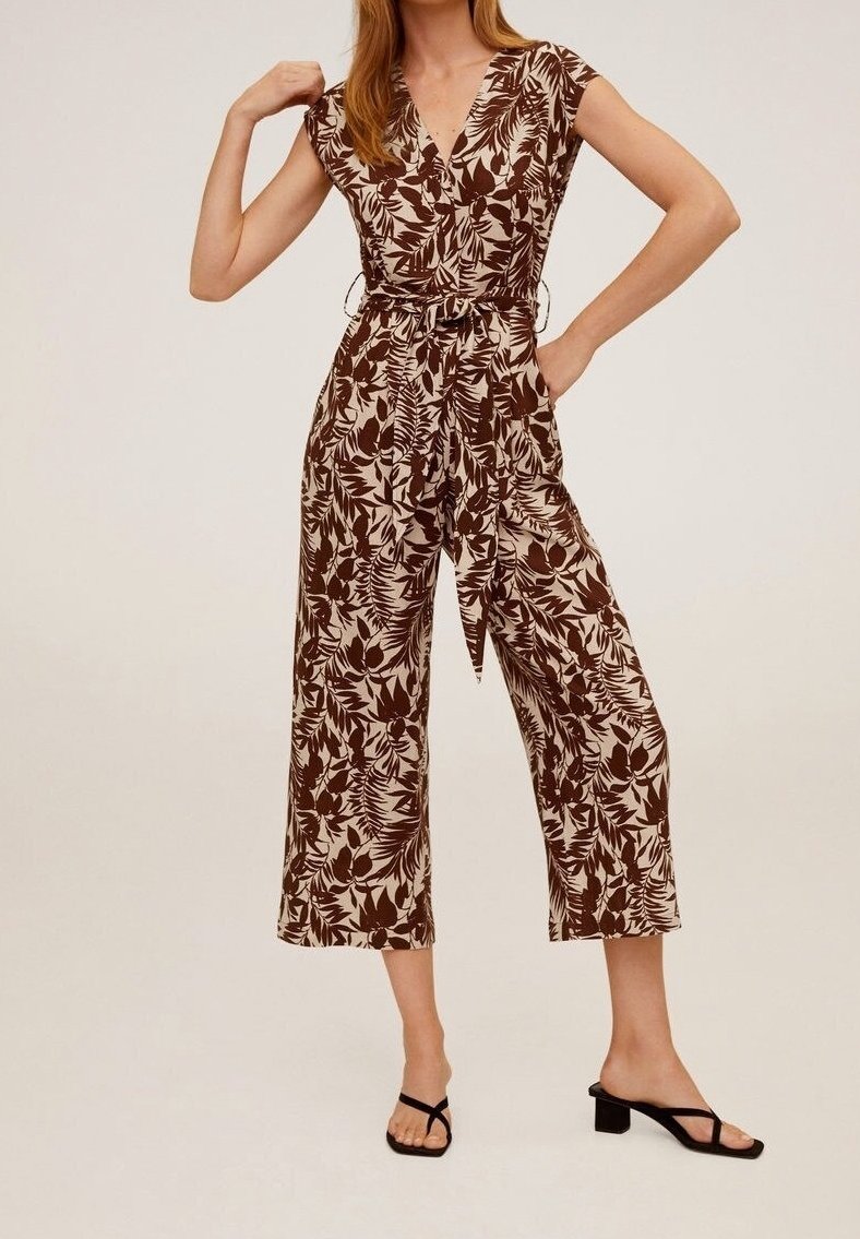 Femme portant une combinaison-short beige et marron à motif floral, manches courtes, jambes larges, ceinture nouée et sandales à talons noires.