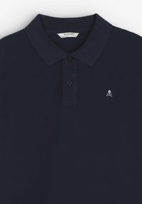 Mörkblå pikétröja i bomullsmaterial, med klassisk krage, treknappsslutning och en liten dödskallelogotyp på bröstet.