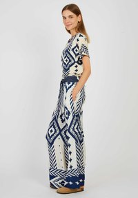 Combinaison à motifs crème avec des designs géométriques bleu marine, manches courtes, jambes larges et une ceinture nouée, avec des poches latérales.