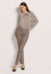 Beige Bluse mit langen Ärmeln und gerafftem Ausschnitt, kombiniert mit körperbetonten braunen Karohosen und schwarzen spitzen High Heels. Glatte Textur überall.