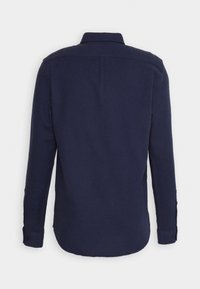 Camisa de manga larga azul marino con puños abotonados, cuello clásico y dobladillo curvado, mostrada desde atrás sobre un fondo liso.