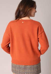 Pull en tricot orange avec un col rond et un ourlet côtelé. Présente des coutures d'épaule tombantes et un petit accent métallique dans le dos.