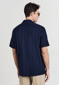 Mann mit Sonnenbrille, navyblauem, kurzärmligem Strukturhemd und beigen Hosen, der leicht abgewandt vor einem schlichten Hintergrund steht.