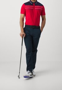 Červená polo triko s námořnickým límcem a pruhovanými akcenty, spárované s námořnickými kalhotami a mnohobarevnými golfovými botami; držící golfovou hůl.