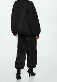 Zwarte oversized jas met een matte textuur, gecombineerd met ruime zwarte broeken. Hoge enkellaarsjes maken de outfit compleet.