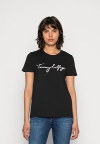 Melnas kokvilnas t-krekls ar īsām piedurknēm, kuram krūtīs ir balts ar rokām rakstīts "Tommy Hilfiger" logo. Apaļa kakla forma.