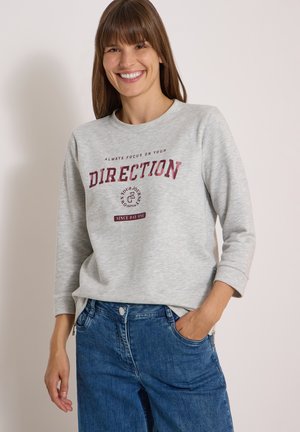 Femme souriante aux cheveux bruns raides porte un sweat-shirt gris avec un texte bordeaux "Always focus on your direction" et un jean bleu, main dans la poche.