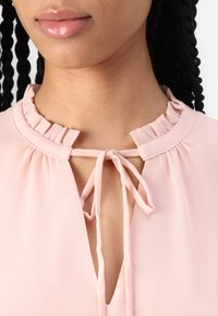 Blusa rosa chiaro con colletto arricciato e chiusura a bow. Presenta un collo a chiave e un tessuto morbido e leggero. Textura liscia.