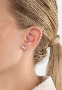 Primo piano di un orecchio di una donna con i capelli biondi raccolti, che indossa un piccolo orecchino a perno a forma di cuore in argento con una pietra centrale.