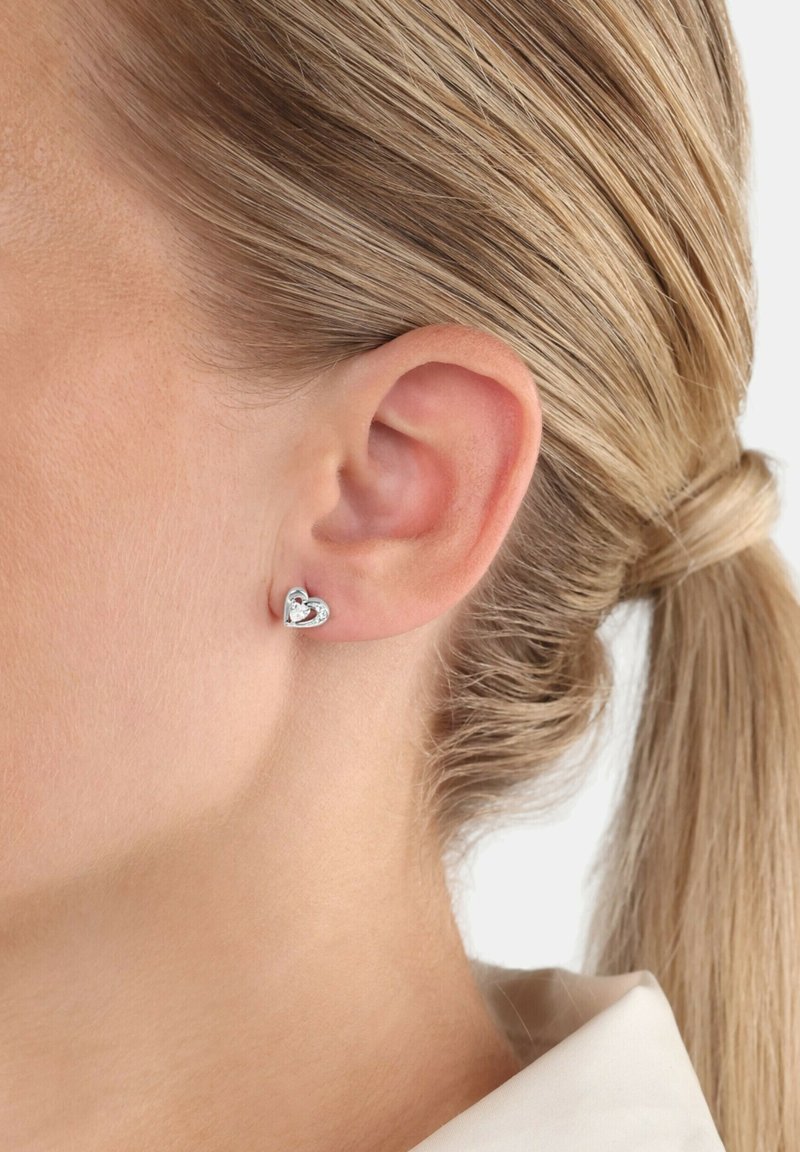 Primo piano di un orecchio di una donna con i capelli biondi raccolti, che indossa un piccolo orecchino a perno a forma di cuore in argento con una pietra centrale.