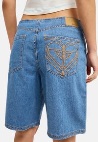 Personne portant un short en jean bleu avec un motif en forme de cœur cousu sur la poche arrière droite et une étiquette "Roxy" sur la ceinture.