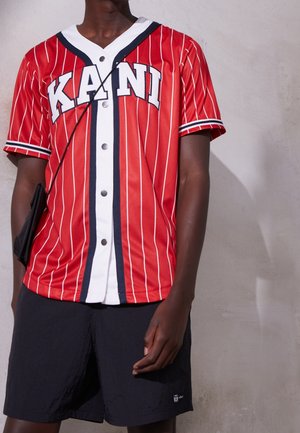 Mężczyzna ubrany w czerwono-białą, paskowaną koszulę na guziki z napisem "KANI", czarne spodenki oraz czarną torbę na ramię.